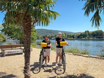 Zwei Radfahrer mit Helmen und gelben Taschen stehen am Sandstrand des Neckarufers in Heidelberg, Palme im Vordergrund, Hügel im Hintergrund.