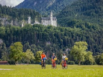 Three cyclists riding through a green meadow with Neuschwanstein Castle visible on a forested hillside in Bavaria.
