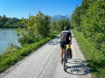 Cyclist with yellow panniers on gravel path along Salzach river with mountains in background, surrounded by green trees under blue sky.