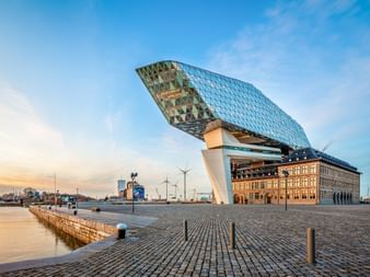 Modernes Glasgebäude mit diamantförmiger Fassade über historischem Hafenmeistergebäude in Antwerpen. Kopfsteinpflasterplatz und Uferpromenade sichtbar.