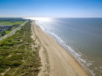 Luftaufnahme des Strandes von Blankenberge in Flandern mit breitem Sandstrand, Dünen, Küstenstraße und ruhiger See unter blauem Himmel.