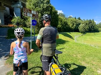 Adult and child cyclists viewing Alpe Adria cycle route signpost in Malborghetto-Valbruna, with green meadows and forest in background.