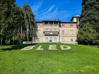 Historisches Gebäude in Bled mit beiger Fassade und dekorativen Balkonen. Weiße Blumen bilden 'BLED' auf dem grünen Rasen. Flaggen und Bäume rahmen die Szene.