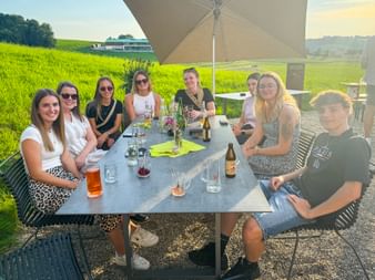 Eight people sitting around outdoor table with drinks and bottles, under parasol with green fields in background during Eurohike summer event.