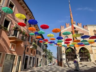 Colorful umbrellas suspended above a street in Soave with historic buildings and a church tower under blue sky.