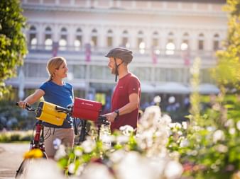 Two cyclists with bikes and panniers pause in a flower garden, with the historic Congress House of Bad Ischl visible in the background.