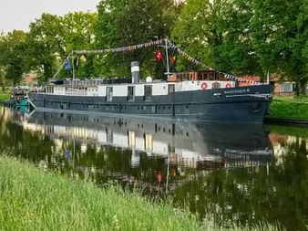 MS Magnifique III, ein Flusskreuzfahrtschiff mit dunklem Rumpf und weißem Oberdeck, liegt an einem ruhigen Kanal. Bunte Flaggen schmücken das Schiff.