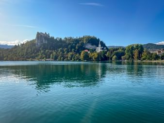 Bleder See mit türkisfarbenem Wasser, Burg Bled auf bewaldeter Klippe und St. Martins Kirche mit weißem Turm. Berge im Hintergrund unter blauem Himmel.