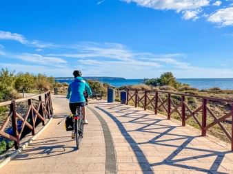 Radfahrer in blauer Jacke auf gepflastertem Küstenweg mit Holzgeländer, Mittelmeer und blauer Himmel im Hintergrund sichtbar.