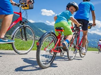 Child in blue helmet and green shorts riding red bike on paved path with adults, green meadows and mountains in background under blue sky.
