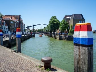Canal view in Dordrecht with drawbridge, historic buildings, and mooring posts painted in Dutch flag colors under clear blue sky.