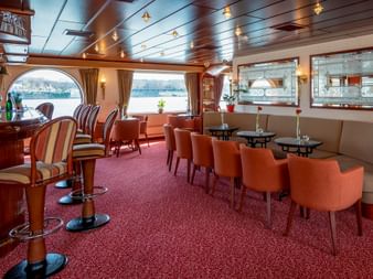 Interior of MS Switzerland bar with orange chairs, round tables, red carpet, and large windows overlooking the water. Wooden ceiling with lights.