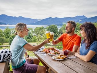 Three cyclists toasting with beer at a wooden table overlooking Lake Tegernsee. Traditional Bavarian snacks are served with mountain views in the background.