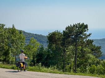 Cyclist with loaded bike on mountain road in Slovenia, overlooking forested hills and the Adriatic Sea in the distance.