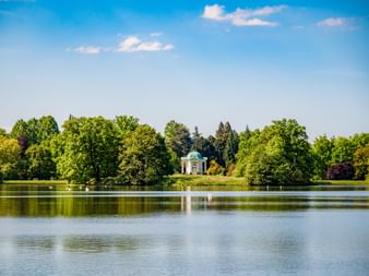 View across a calm lake reflecting trees and sky, with a small turquoise-domed pavilion surrounded by lush green trees in Kassel.