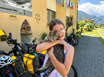 Smiling child with glasses holding a tabby cat in Chiusaforte. Bicycles with yellow bags and mountains visible in background.