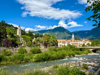 Meran cityscape with Passer River flowing through green vegetation, historic church towers, and mountain backdrop under blue sky.