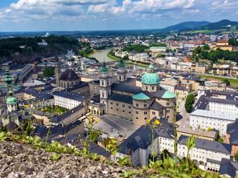 Panoramic view of Salzburg's old town with cathedral dome, historic buildings, and Salzach River. Mountains visible in the background under blue sky.