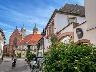 Radfahrer auf Kopfsteinpflasterplatz in Ladenburg mit historischer Kirche, Fachwerkhäusern und Straßencafé unter blauem Himmel.