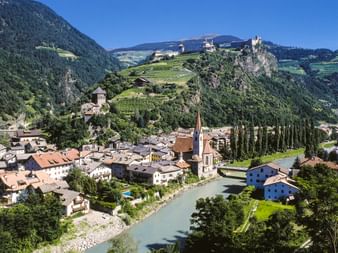 Alpine town of Klausen with church spire along a river, surrounded by mountains with castles and monastery on hilltops under blue sky.