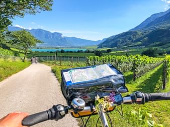 Fahrradlenker mit Kartenhalter auf Radweg durch Apfelplantagen, Berge und See im Hintergrund bei Bozen.