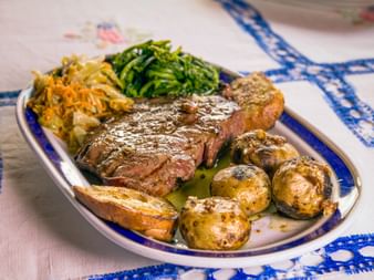 Gegrilltes Posta Mirandesa Rindersteak auf weißem Teller mit Röstkartoffeln, sautiertem Grün und Gemüse auf blau karierter Tischdecke.