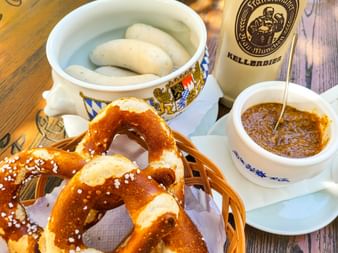 Traditional Bavarian breakfast with white sausages in a decorative bowl, soft pretzel with salt, sweet mustard, and Kellerbier bottle on wooden table.