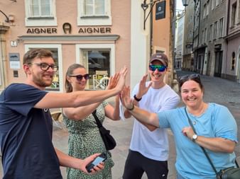 Four people doing a group high-five on a cobblestone street in front of historic buildings with an Aigner store visible in the background.