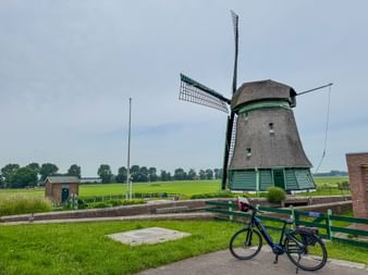 Blue rental bicycle parked on path in front of traditional Dutch windmill with green trim. Green fields and trees visible in background under cloudy sky.