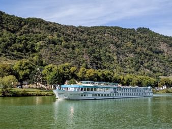 MS Switzerland river cruise ship sailing on calm green water with forested hills in the background under a partly cloudy sky.