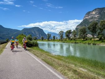 Three cyclists with yellow panniers riding along the Etsch cycle path beside a turquoise river, with dramatic mountains in the background.