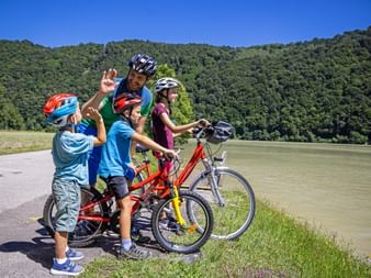 Family of four with bicycles giving high-fives along the Lech River. Green hills and blue sky in background, river visible beside path.