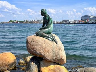 Bronze statue of The Little Mermaid sitting on a rock in Copenhagen harbor with blue water and city skyline in the background.