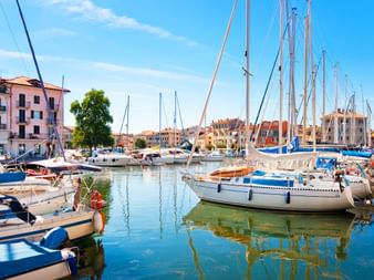 Hafen in Grado mit zahlreichen Segelbooten im ruhigen Wasser. Bunte Gebäude säumen die Uferpromenade unter blauem Himmel mit leichten Wolken.