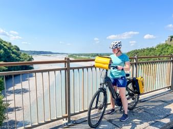 Radfahrerin mit weißem Helm und gelben Taschen auf Brücke über den Tagliamento in Friaul, mit breitem Flussbett und grünen Hügeln.