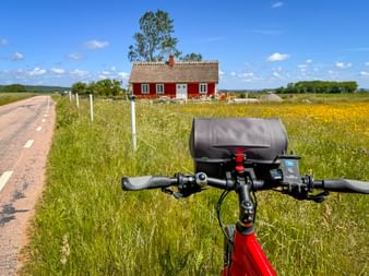 Fahrradlenker mit Lenkertasche vor einem traditionellen roten Schwedenhaus mit weißen Akzenten, umgeben von gelben Wildblumen und grünen Feldern.