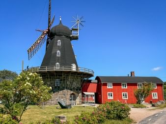 Dunkle Holzwindmühle mit Flügeln neben einem roten schwedischen Bauernhaus mit weißen Akzenten, umgeben von grünem Rasen und Blumen.