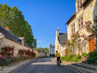 Cyclist in yellow jacket riding through a quiet Provence village street with stone houses, climbing roses, and blue sky.
