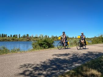 Two cyclists with yellow panniers riding on a paved path beside a river with cypress trees in the background under clear blue sky.