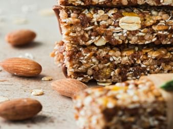 Stack of homemade muesli bars with oats, almonds, and seeds on a wooden surface. Loose almonds are scattered beside the bars.