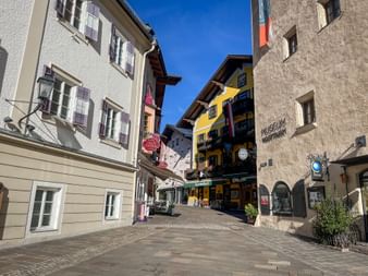 Cobblestone street in Zell am See lined with colorful historic buildings. The Vogtturm tower is visible on the right under a clear blue sky.