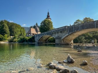 Historic stone bridge spanning clear water at Lake Bohinj with a church tower and mountains in the background under blue sky.