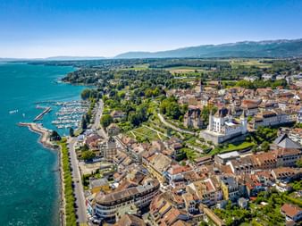 Luftpanorama von Nyon am Genfersee mit historischem Stadtkern, Schloss, Hafen und umliegenden Bergen unter klarem blauem Himmel.