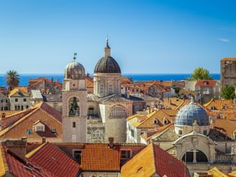 Aerial view of Dubrovnik's old town with terracotta-tiled roofs, church domes, and bell towers against the blue Adriatic Sea.