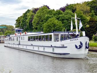 Weißes Flusskreuzfahrtschiff MS Fluvius mit blauem Zierstreifen liegt an einem Kanal, umgeben von üppigen grünen Bäumen unter bewölktem Himmel.
