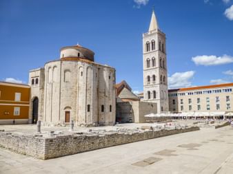 St. Donatus Church with circular stone structure and tall bell tower in Zadar, Croatia. Ancient Roman ruins visible in foreground plaza.
