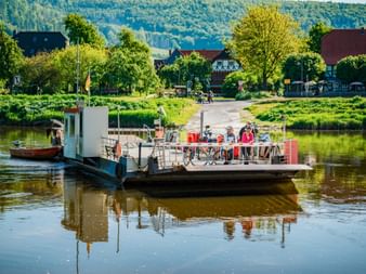 Radfahrer mit Rädern auf einer Fähre über die Weser bei Hemeln. Traditionelle Fachwerkhäuser und grüne Hügel im Hintergrund sichtbar.