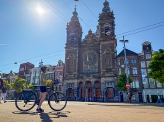 Radfahrer mit Fahrrad vor der Nikolauskirche in Amsterdam. Die barocke Kirche mit Zwillingstürmen und Uhr steht am Prins Hendrikkade Platz.