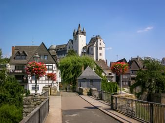 Historische Brücke zum Grafenschloss Diez mit weißen Türmen. Traditionelle Fachwerkhäuser mit roten Blumenkästen säumen das Flussufer.