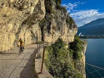 Cyclist in yellow jersey on scenic Strada del Ponale path carved into cliff above Lake Garda, with mountains and blue sky in background.